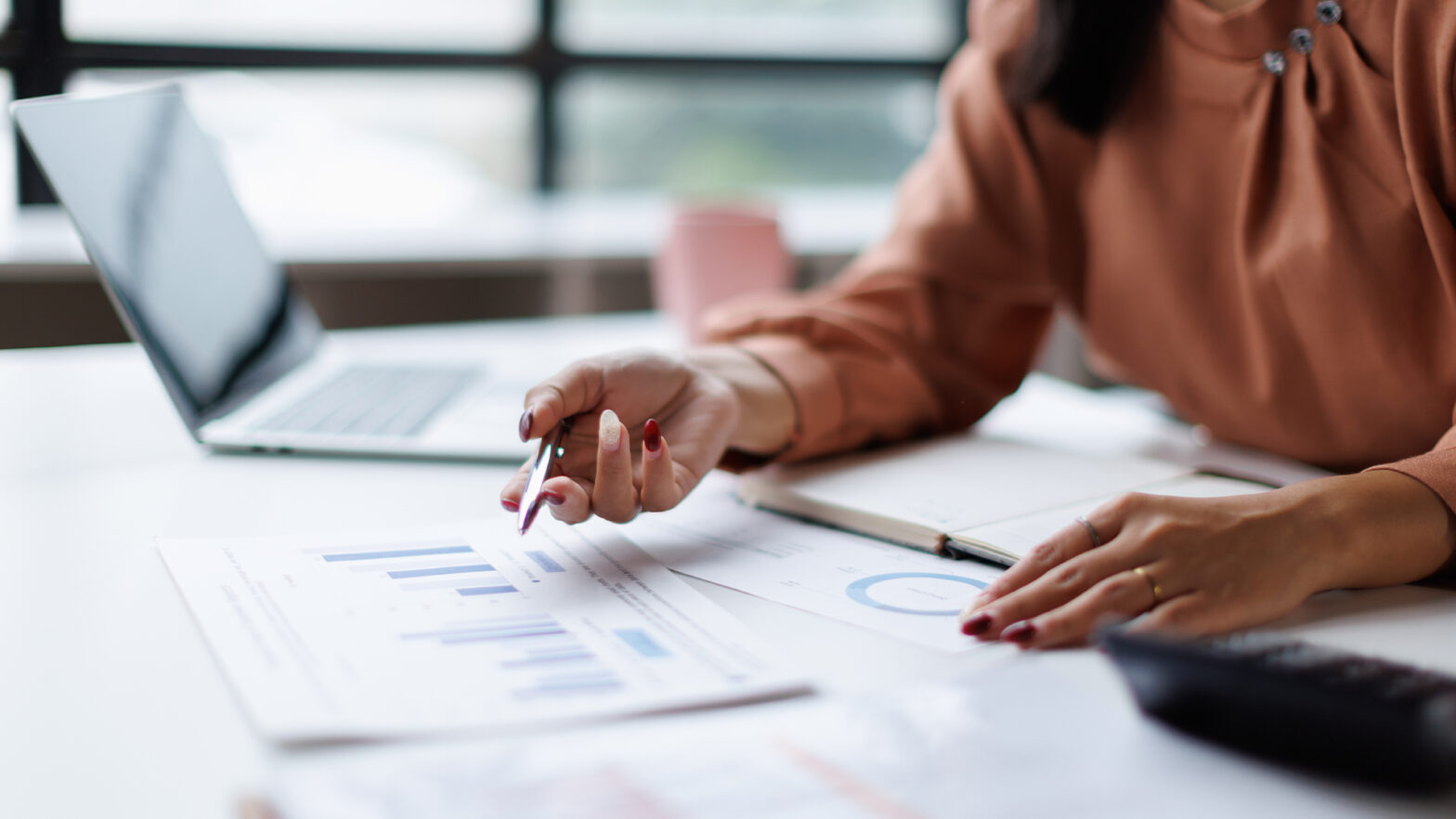 Professional woman analyzing financial data and business reports, using a pen to point at charts while working on a laptop at an office desk